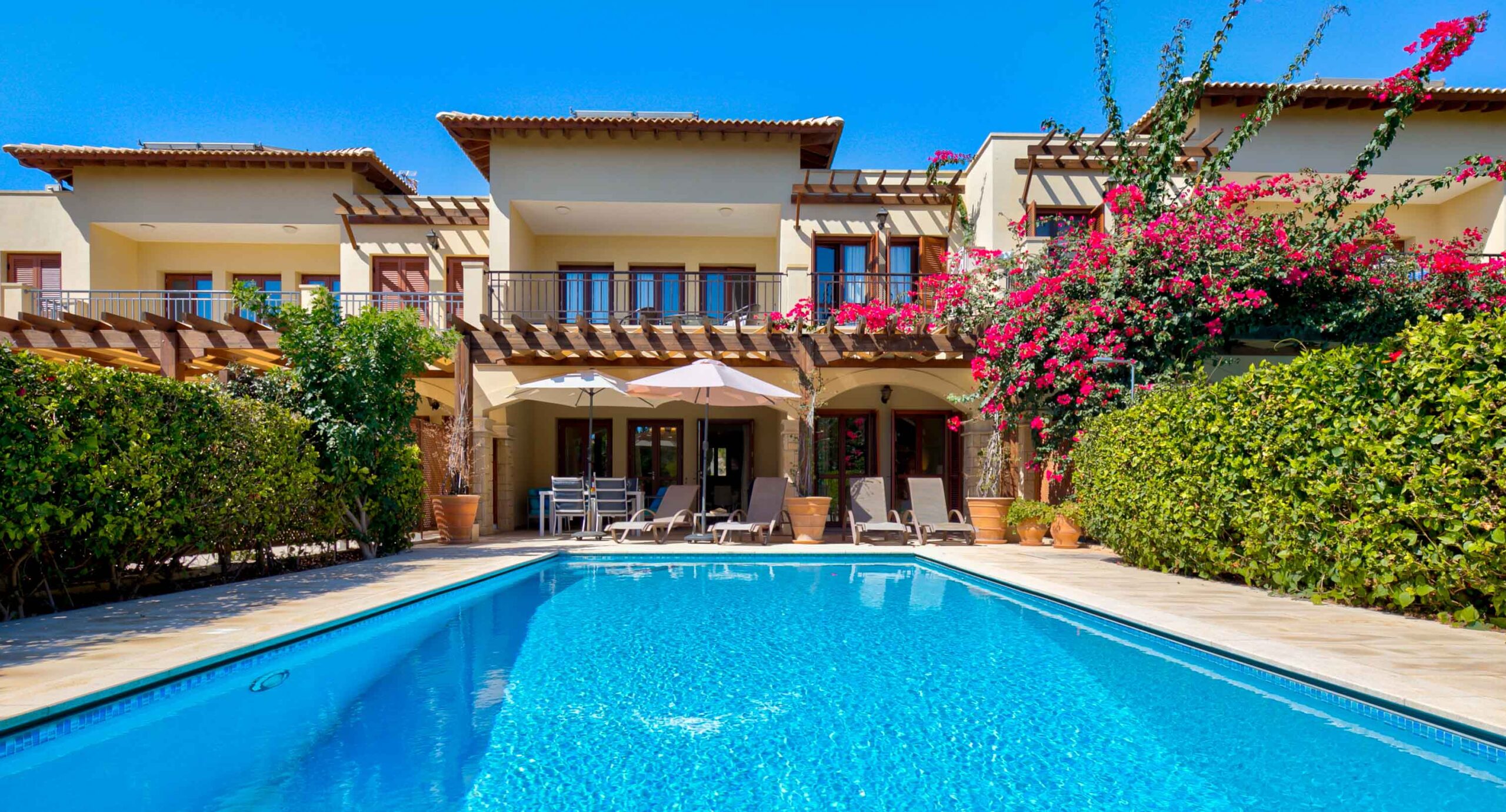 Looking from the far end of a rectangular private pool to the apartment building of Apartment AT02 on Aphrodite Hills Resort, showing sunbeds and parasols on the apartment terrace near the ground floor doors. High hedges line the terrace either side.