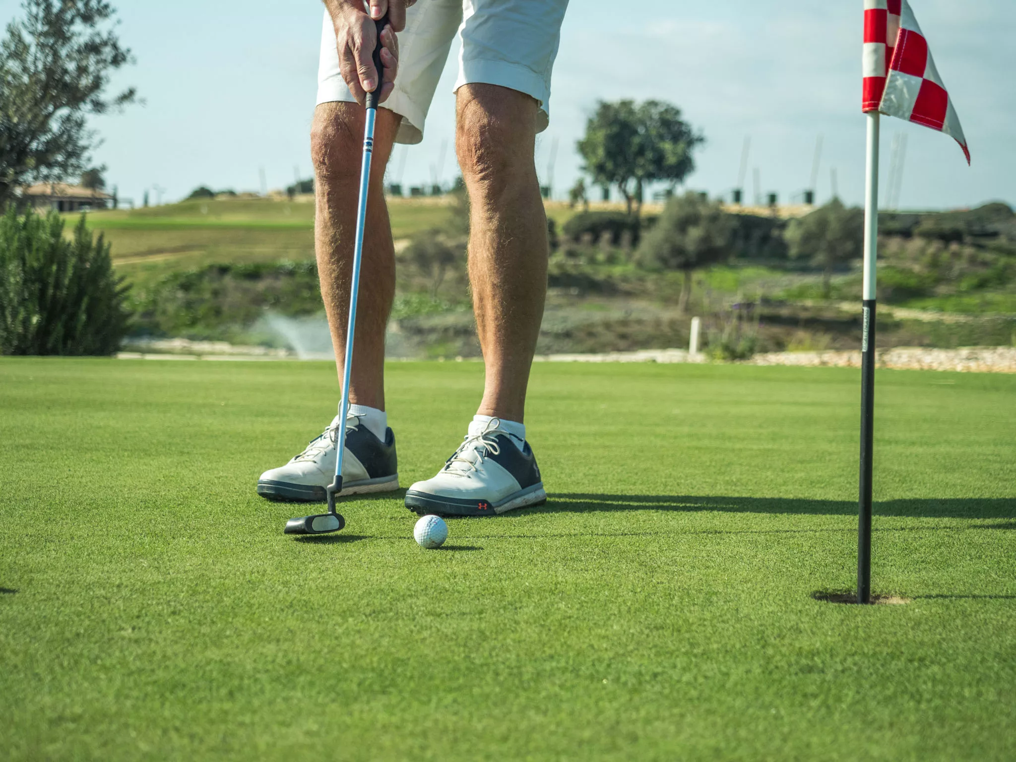 Man's legs and putter as he is poised to putt the ball into golf hole, with practice facilities at Aphrodite Hills PGA National Golf Course in background.