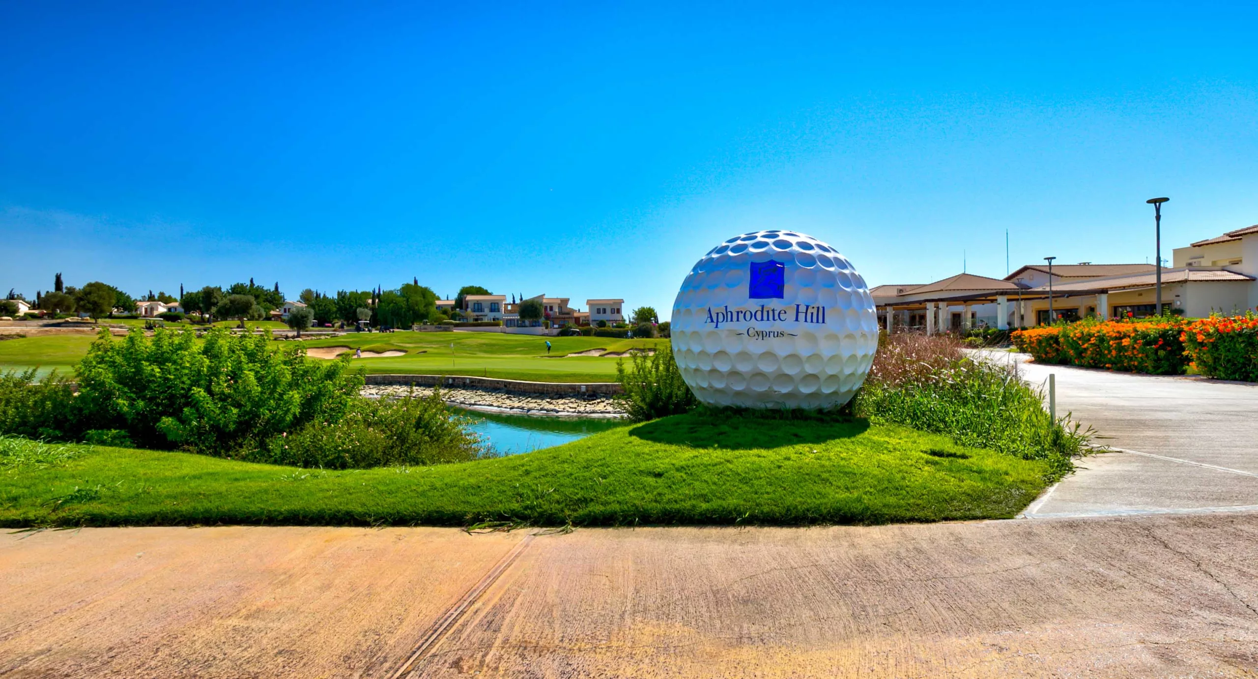 View of Aphrodite Hills Golf Clubhouse and 18th green from the chipping area, with an oversized golf ball in the foreground.