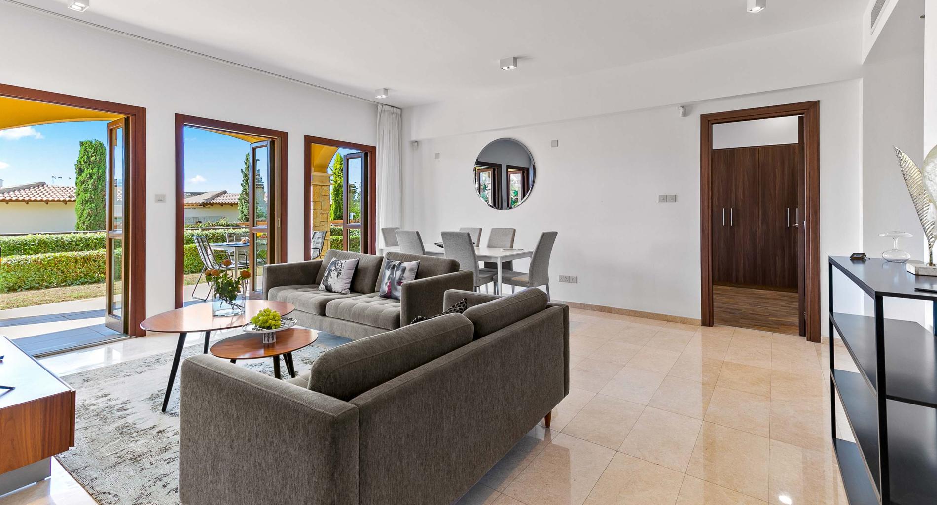 Bright living and dining room, white walls, comfortable brown sofas and grey carpet, with marble floors. Doors opening to private garden and outside dining furniture. Apartment AP02 on Aphrodite Hills Resort.