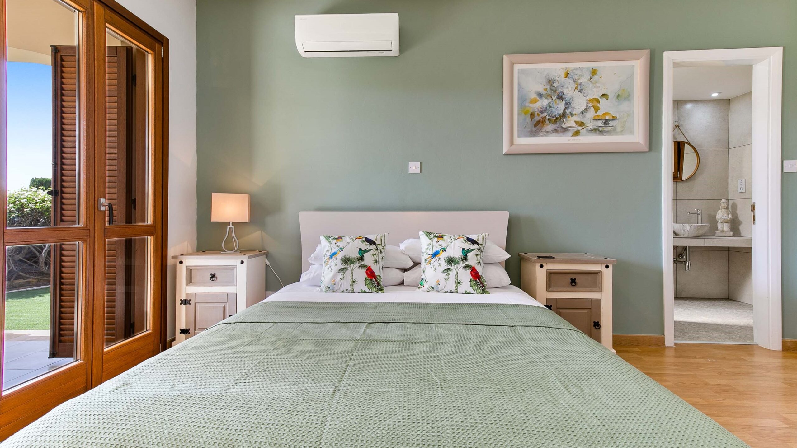 double bed with light green bedding, against light green wall, with paining and air con unit on the wall. Bedroom of Apartment Eleos on Aphrodite Hills Resort.