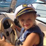 Little girl holding a boat wheel, wearing a captain's hat, smiling in the sunshine.
