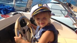 Little girl holding a boat wheel, wearing a captain's hat, smiling in the sunshine.