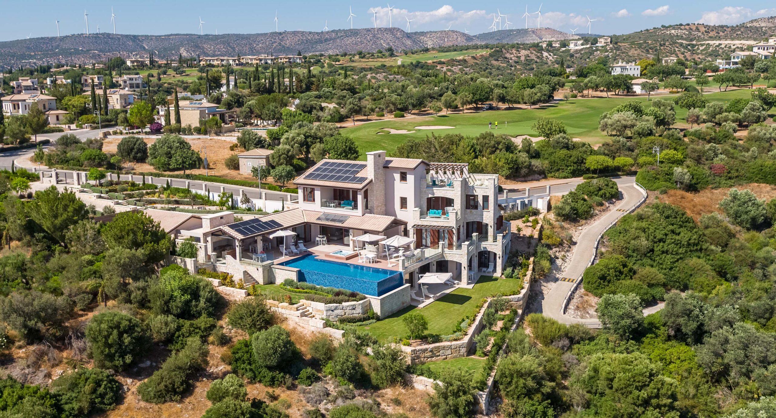 Aerial view of Villa 102 on Aphrodite Hills, facing the front with pool, terraces, whole villa building, surrounding ground in view. Back drop shows golf course and mountain views.