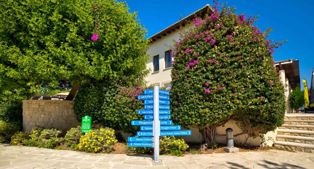 Blue sign posts indicating various facilities at Aphrodite Hills Resort, placed in front of lush trees and bougenvelia bush, in Aphrodite Hills Resort village centre.