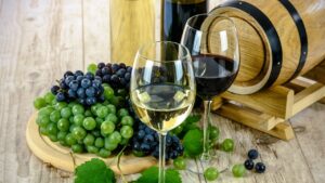 Two wine glasses, one with red one with white wine, placed infront of a bunch of grapes and a wine barrel.